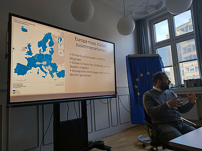 Johannes Schraps an einem Tisch vor einem Active-Panel. Auf dem Panel folgende Folie: Überschrift: Europa muss stärker zusammenarbeiten. Links eine Europakarte mit blau markierten Nato-Staaten, grau markierten Nicht-Nato-Staaten und grün markierten demilitarisierten Gebieten. Stand der Karte ist März 2024. Zusatzangabe in der Legende: “The special status of the Åland Islands as demilitarised area is not affected by Finland’s membership in NATO.” Rechts Stichpunkte: Finnland und Schweden sind NATO-Mitglieder; Ostsee ist heute fast vollständig von NATO-Staaten umgeben; Strategischer Vorteil gegenüber Russland deutlich gewachsen.