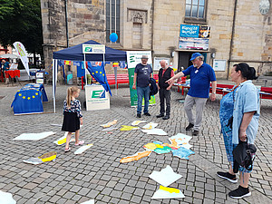 Im Hintergrund die Fassade der Marktkirche St. Nicolai, Hameln. Davor ein Zeltdach der Europa-Union, daran eine Wimpelkette mit Flaggen europäischer Länder, sowie eine Europaflagge. Vor dem Dach zwei Aufsteller der EUD. Links vom Dach ein Tisch mit der Europaflagge als Tischdecke, darauf Promomaterial. Im Vordergrund ein hölzernes Europapuzzle, sowie vier Erwachsene rechts (davon ein EUD-Mitglied mit europablauem Shirt und Kappe) und links ein Kind.